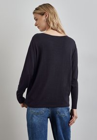Pull tricot bleu marine à coupe ample, décolleté arrière bas et poignets côtelés, associé à un jean bleu clair. Texture douce, design décontracté.