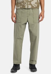 Green cotton trousers with a relaxed fit, featuring two front pockets and topstitching details. Worn with black sneakers.