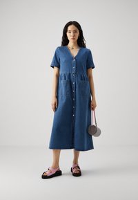 Denim maksi haljina s kratkim rukavima, prednjim dugmadi i dva prednja džepa. Uparena s ružičastim platformama i okruglom torbicom od perlica.