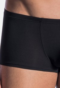 Black swim trunks made of smooth, stretchy material feature a fitted design, short length, and flatlock seams for comfort.