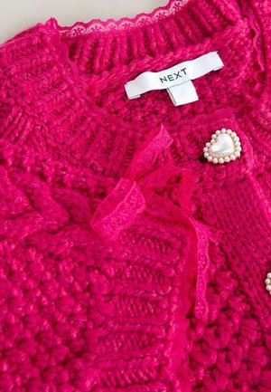 Lyserød strikket cardigan med ribbet struktur, blonderkant og dekorativ hjerteformet brosje prydet med perler. Mærketiketten er synlig.