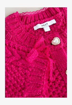 Hellrosa gestrickte Strickjacke mit gerippter Struktur, Spitzenbesatz und dekorativer herzförmiger Brosche, verziert mit Perlen. Markenetikett sichtbar.