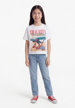 Jeune fille souriante, portant un T-shirt blanc avec un requin en talons rouges imprimé et un jean bleu, debout devant un fond blanc.