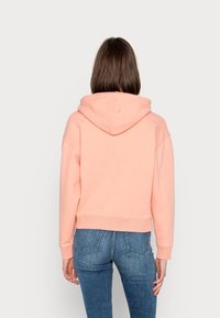 Jasnoróżowa krótka bluza z miękkiego materiału, z dużym kapturem i prążkowanymi mankietami, zestawiona z niebieskimi dżinsami. Prosty design, bez dodatkowych elementów.