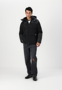 Sort puffers jakke med aftagelig hætte, lynlås foran og elastiske manchetter. Båret sammen med mørke jeans med et knæhul og sorte støvler.