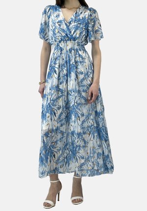 Kobieta w niebiesko-białej kwiatowej sukience midi z bufiastymi rękawami, marszczoną talią i białymi sandałami na wysokim obcasie.