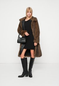 Blonda sieviete, kas valkā brūnu kažokādu, melnus kleitas, melnas augstas zābakus un tur melnu ādas rokassomu uz baltā fona.