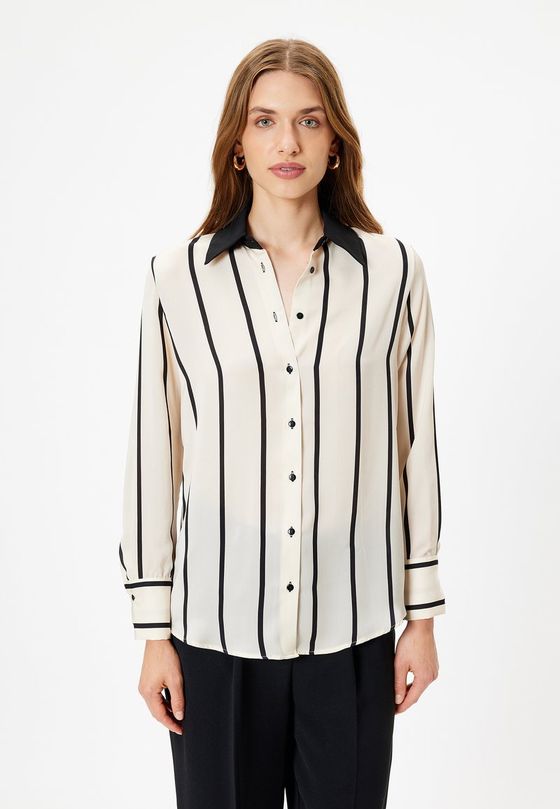 Crème blouse met verticale zwarte strepen, lange mouwen en een zwarte kraag. Voorzien van knopen aan de voorkant en polsmanchetten met bijpassende zwarte strepen.