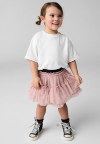 T-shirt bianca in cotone, gonna in tulle rosa con volants a strati e scarpe alte nere con lacci bianchi e dettagli stampati.