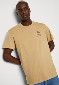 Beige cotton t-shirt with short sleeves, round neckline, and a small graphic print of a tree and text on the left chest.