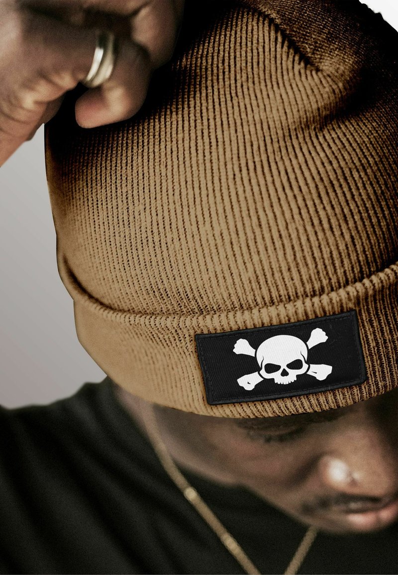 Brown ribbed beanie made of knit fabric featuring a black patch with a white skull and crossbones design. No other notable details.