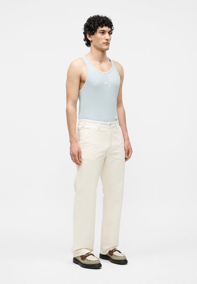 Νέος άνδρας με σγουρά μαλλιά, που φοράει ανοιχτό μπλε ριμπ μπλουζάκι, παντελόνι σε εκρού χρώμα και δίχρωμα loafers, στέκεται μπροστά σε ένα μονόχρωμο φόντο.