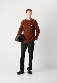 Bruine geribde trui met een klein logo-patch, gecombineerd met donkere spijkerbroek en zwarte schoenen. Model houdt een zwarte tas vast. Eenvoudig, modern ontwerp.