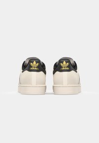 Adidas sneakers with a cream upper, black heel cap, yellow logo, and textured detailing. Flat rubber sole and rounded toe design.