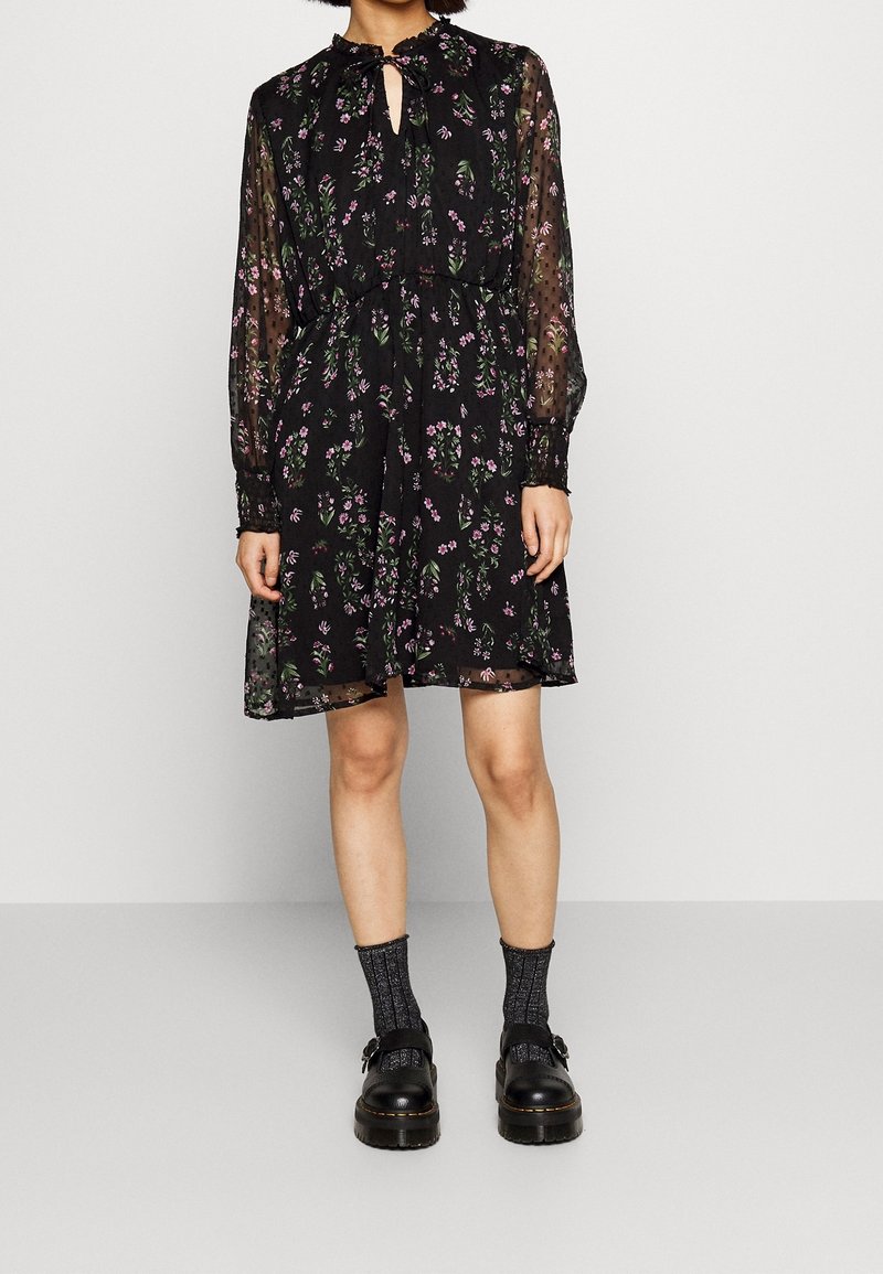 Vestido negro con estampado floral, mangas largas transparentes, escote con abertura en forma de llave, cintura con cordón y falda fluida. Llevado con zapatos de plataforma negros y calcetines.