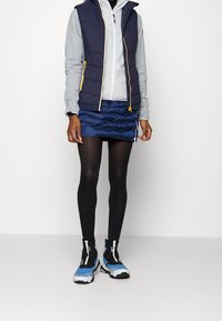 Marinblå puffer-väst över en ljusgrå hoodie, tillsammans med en marinblå kviltad minikjol och svarta leggings. Inkluderar blå och svarta högstövlar.