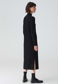 Abito lungo a maniche lunghe nero a coste con colletto alto e spacchi laterali. Tessuto morbido, silhouette aderente, cade sotto le ginocchia. Abbinato a scarpe e calze nere.