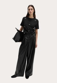 Černé tričko s malými ozdobami v podobě mašliček, v kombinaci s volnými, vysoce lesklými černými kalhotami. Modelka drží lesklou kabelku v odpovídající barvě.