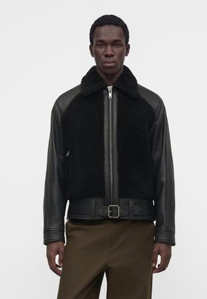 Homme portant une veste en fourrure noire et en cuir avec fermeture éclair et ceinture, associée à un pantalon marron, debout devant un fond blanc uni.