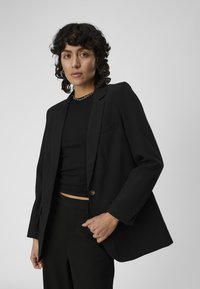 Blazer nero realizzato in tessuto liscio, con chiusura a un bottone, rever a scollo e vestibilità comoda, indossato sopra un top nero a costine.