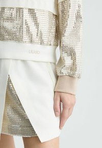Beige and gold sequined top with a white skirt, featuring a textured design, elastic cuffs, and a logo on the hem.
