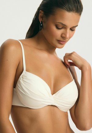 Žena v bielom podprsenke s tenkými ramienkami, s čiernymi kruhovými náušnicami, hľadí dole a dotýka sa ramienka podprsenky na ramene.