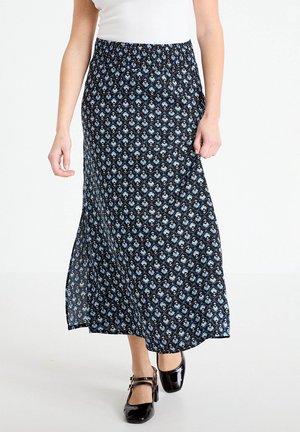 Kobieta ubrana w czarną spódnicę midi z niebieskim kwiatowym wzorem, białą bluzkę i czarne lakierowane szpilki z paseczkami na kostkę, idąca do przodu.