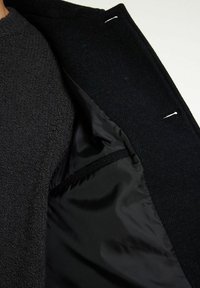 Mörk yllekappa med ett mjukt, texturerat ytterlager och en slät, svart satinfoder. Inkluderar en ficka och vita bastrådar.