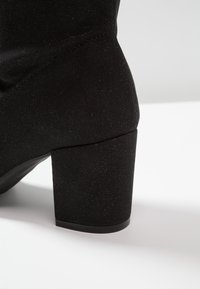 Botín de tobillo de tela negra que presenta un upper texturizado con un sutil brillo, un tacón bloque robusto y detalles de costura sin costuras.
