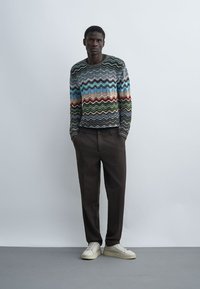 Un homme est appuyé contre un mur uni, portant un pull à rayures ondulées multicolores, un pantalon foncé, des baskets blanches, les mains dans les poches, expression neutre.