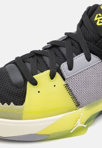 Gros plan d'une chaussure de sport noire, grise et jaune avec des lacets noirs, un tissu en mesh et un logo Jumpman blanc sur une semelle translucide jaune.