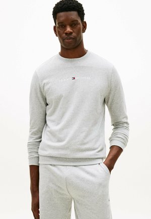 Czarnoskóry mężczyzna w jasnoszarej bluzie Tommy Hilfiger i dopasowanych spodniach dresowych, stojący z jedną ręką w kieszeni.