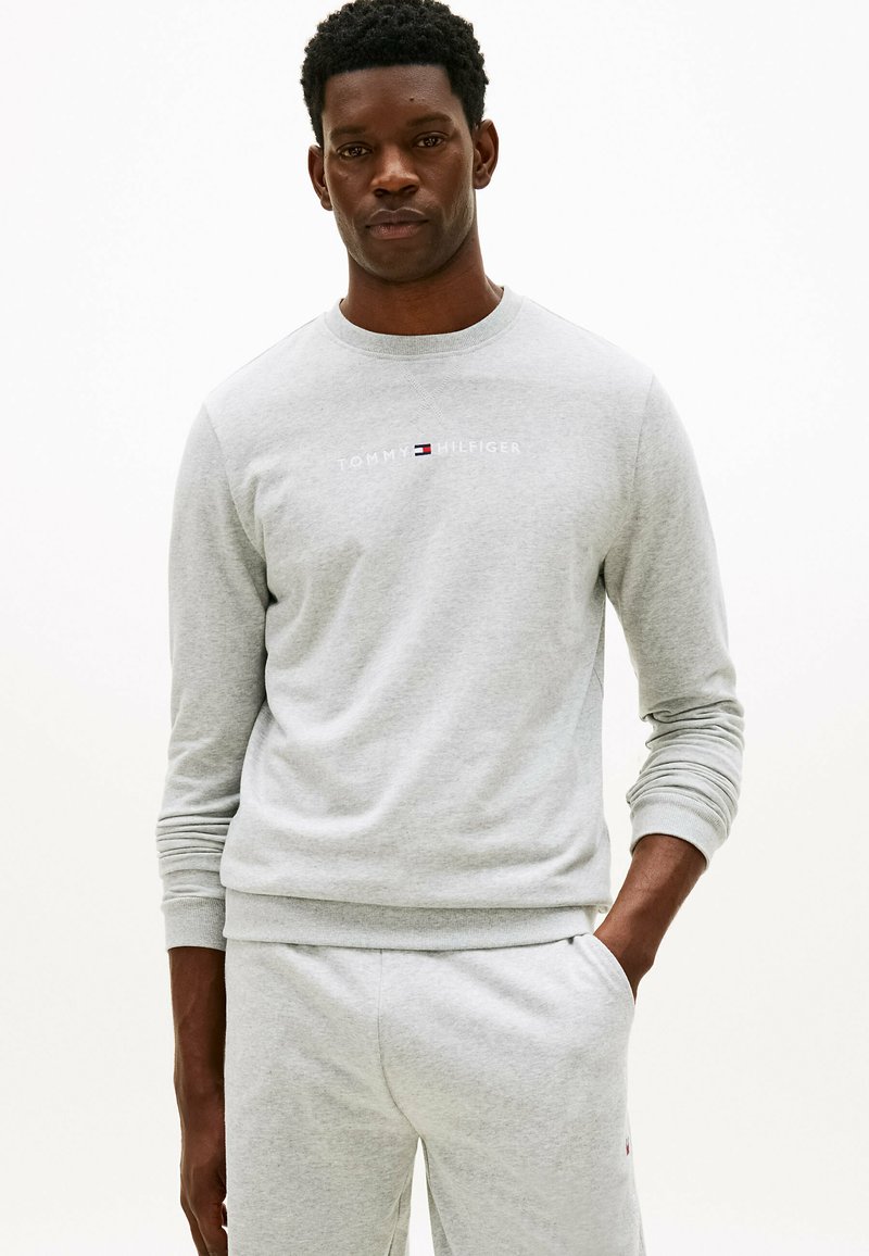 Schwarzer Mann trägt ein hellgraues Tommy Hilfiger Sweatshirt und passende Jogginghose, steht mit einer Hand in der Tasche.