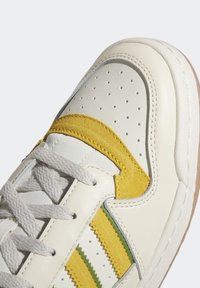 Zapatillas de cuero blanco con acentos de ante amarillo y verde, puntera perforada, cordones grises y una suela de goma marrón.