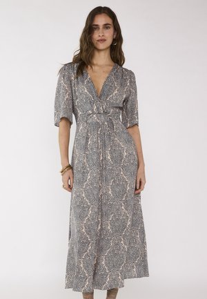 Vrouw met golvend bruin haar draagt een grijs-beige midi-jurk met V-hals en patroon, ellebooglange mouwen, gouden armbanden en oorringen.