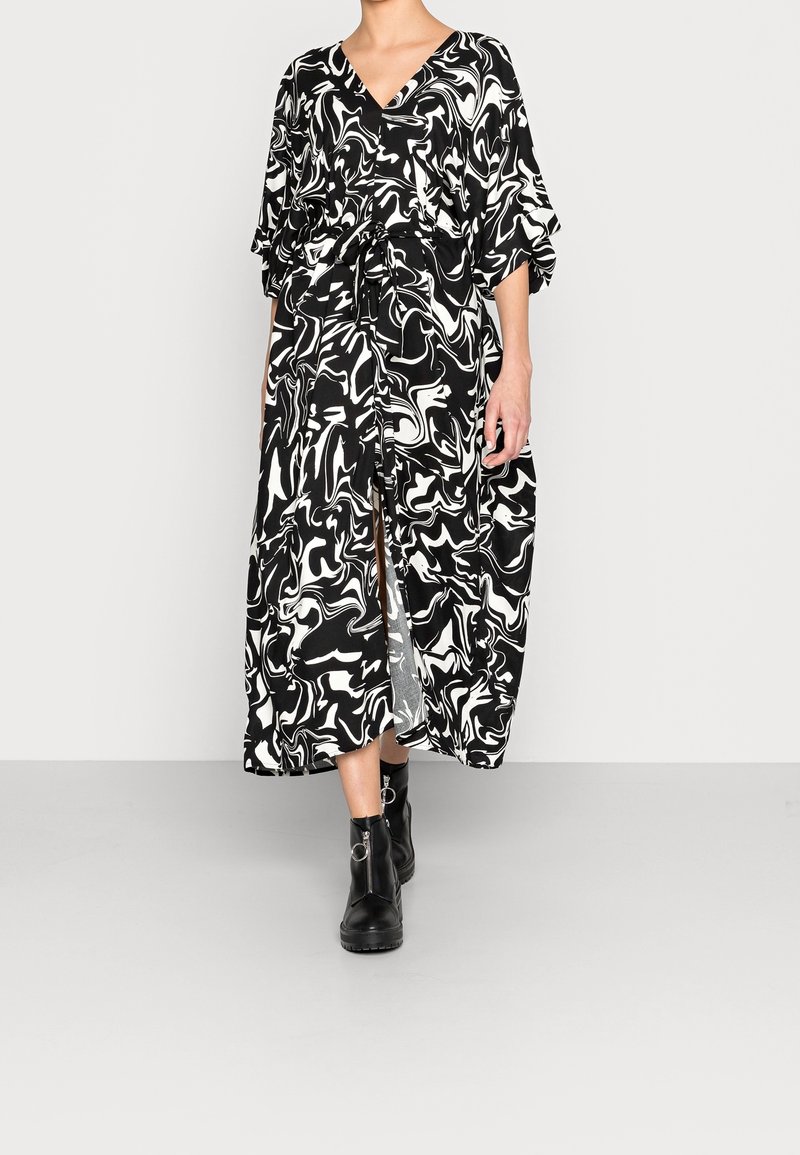 Robe maxi à motif marbre noir et blanc, avec un col en V, des manches courtes et une taille nouée. Fentes notables de chaque côté. Portée avec des bottes noires.