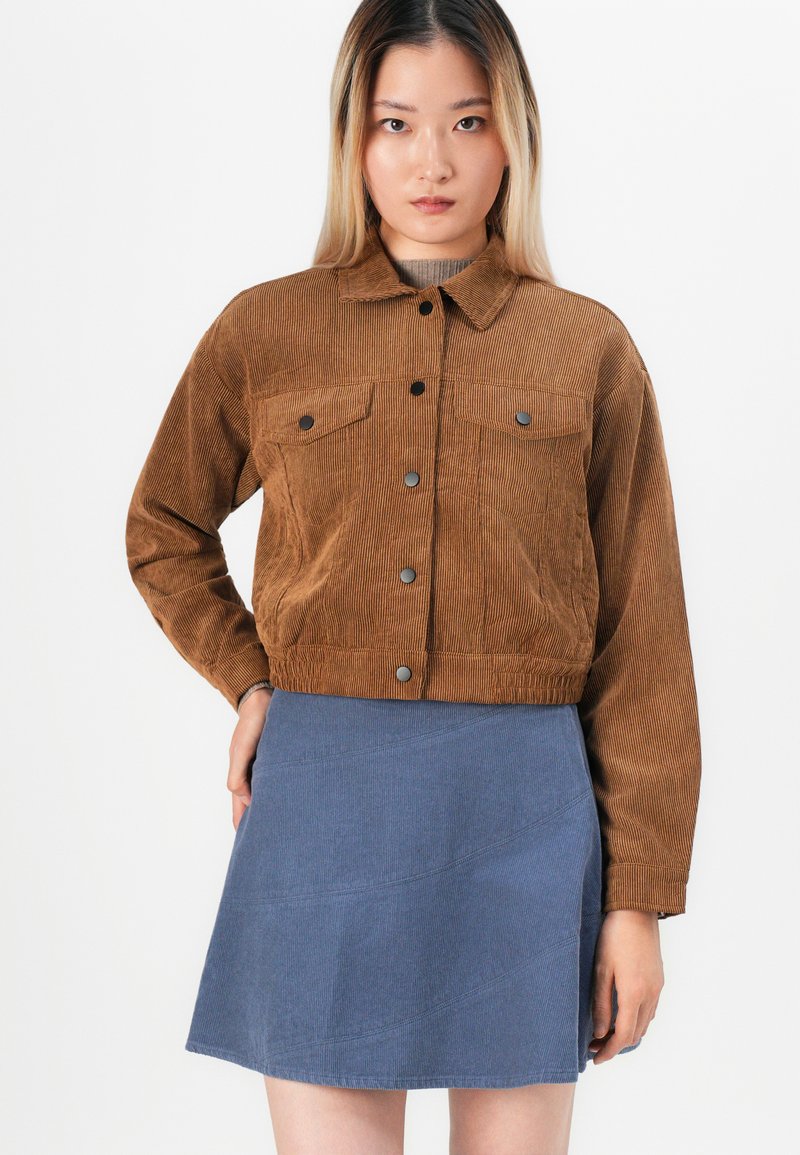 Jeune femme portant une veste en velours côtelé marron avec des boutons noirs et une jupe en velours côtelé bleue, se tenant devant un fond blanc uni.