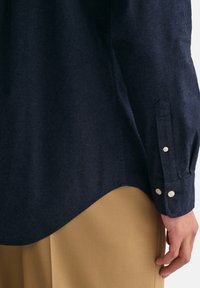 Donkerblauwe shirt in een gestructureerde stof, met lange mouwen met knoopmanchetten en een gebogen zoom, gecombineerd met beige broeken.