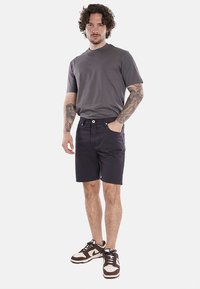 T-shirt grigia a maniche corte abbinata a pantaloni corti neri; modello che indossa scarpe da ginnastica marroni e bianche; tatuaggi visibili sulle braccia.