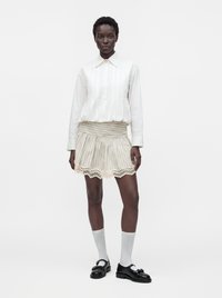 Femme portant un chemisier blanc à col, une jupe beige rayée à volants avec bordure en dentelle, des chaussettes blanches et des mocassins noirs, debout devant un fond uni.