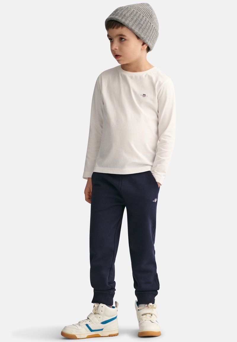 Langermet hvit bomullsskjorte, marineblå joggebukser og hvite høye sneakers med blå detaljer. Barn som har på seg en grå strikket lue.
