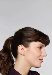 Kolczyki wkrętki ze stali chirurgicznej z niebieskim kamieniem szlachetnym, w kształcie okrągłym, o polerowanym wykończeniu. Noszone przez osobę z ciemnobrązowymi włosami.