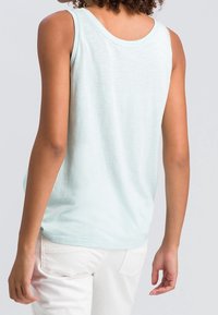 Mujer vista desde atrás que lleva una camiseta sin mangas de color azul claro y pantalones blancos, contra un fondo gris liso.