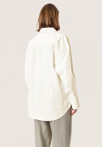 Hvid oversized skjorte med rygsøm, lange ærmer og buet kant. Teksturstof med en glat overflade. Kombination med grå bukser.