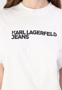 Vit bomulls-T-shirt med fett svart tryck som lyder "KARL LAGERFELD JEANS" över bröstet. Klassisk rund halsringning.