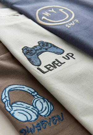 Trois T-shirts avec des motifs brodés : une manette de jeu bleue, des écouteurs et du texte. Les couleurs incluent le gris clair, le bleu foncé et le marron.