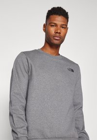 Man in een grijze langekous crewneck sweatshirt met het The North Face-logo op de linkerkant van de borst, staand tegen een effen lichte achtergrond.