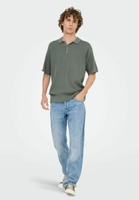 Oliven grøn strikket polo shirt med korte ærmer, 3-knaps lukning, parret med lyseblå straight-leg jeans og hvide sneakers med brune detaljer.