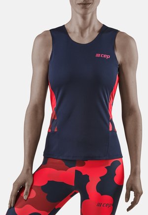 Kobieta ubrana w granatowy sportowy top bez rękawów z różowym logo oraz pasujące czerwono-granatowe legginsy w wzór moro, stoi ze zrelaksowanymi rękami.