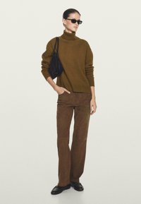 Maglione a collo alto marrone, tessuto morbido, abbinato a pantaloni scamosciati marroni, entrambi slim fit. Gli accessori includono una borsa nera e occhiali da sole scuri.